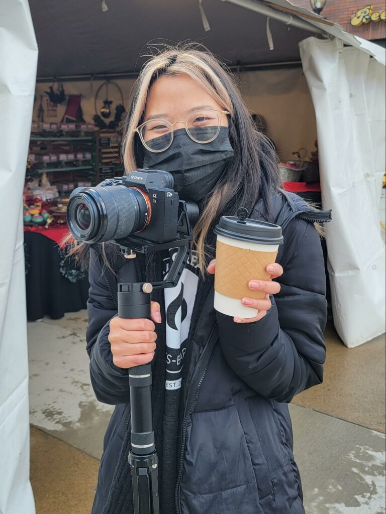 Coal Creative’s Video Specialist Jay Nguyen shows off the essentials — a camera and a hot beverage — at the Holiday Market at Midtown Village on November 27.